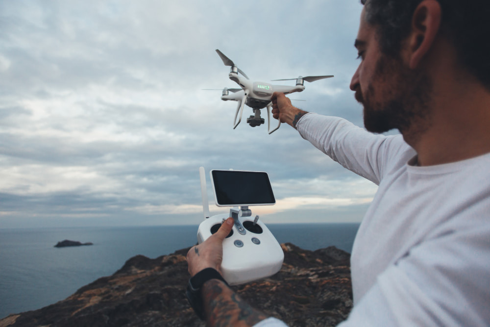 A person with a beard, in a white shirt, controls a drone with a remote over a rocky coastline.