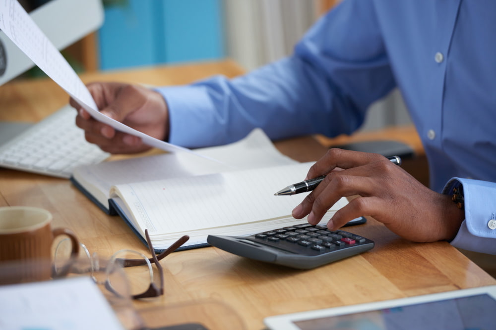 Person in blue shirt uses calculator with paper, notebook, coffee, and computer—showing mortgage focus and productivity.