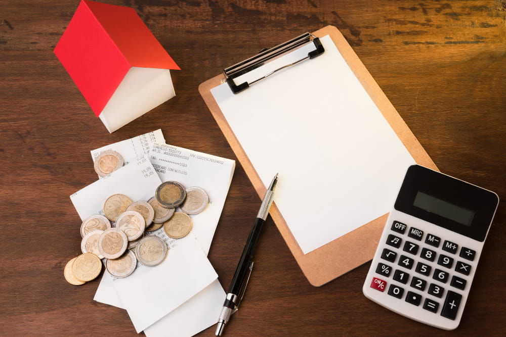 Red-roofed house model, coins, receipts, clipboard, pen, and calculator on wood—symbolizing mortgage financial planning.
