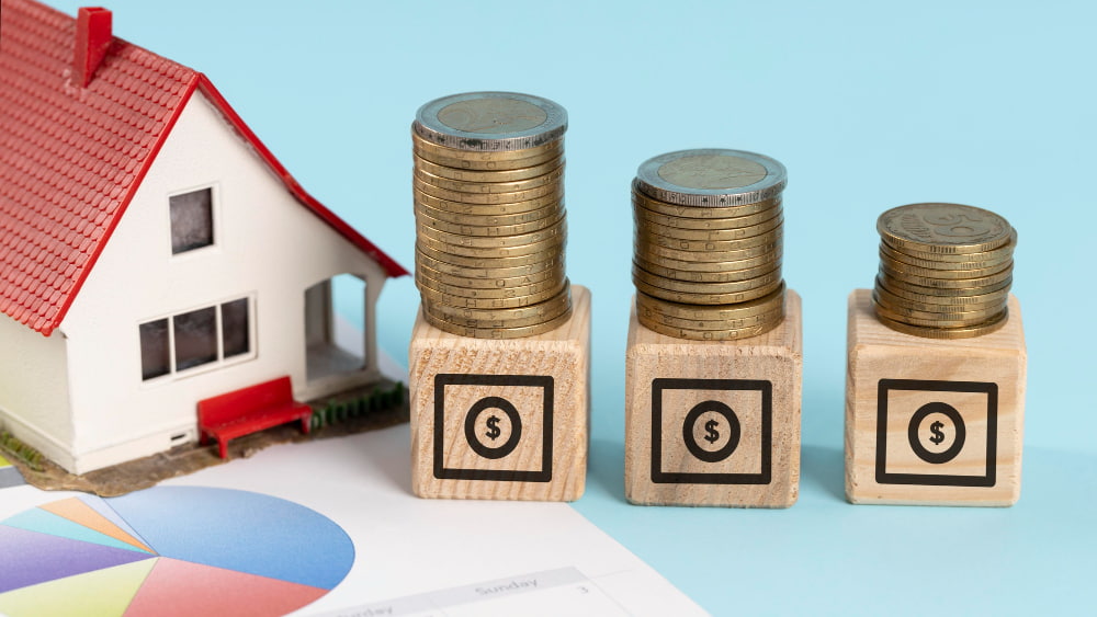 A model house with a red roof sits beside stacks of coins on wooden blocks.