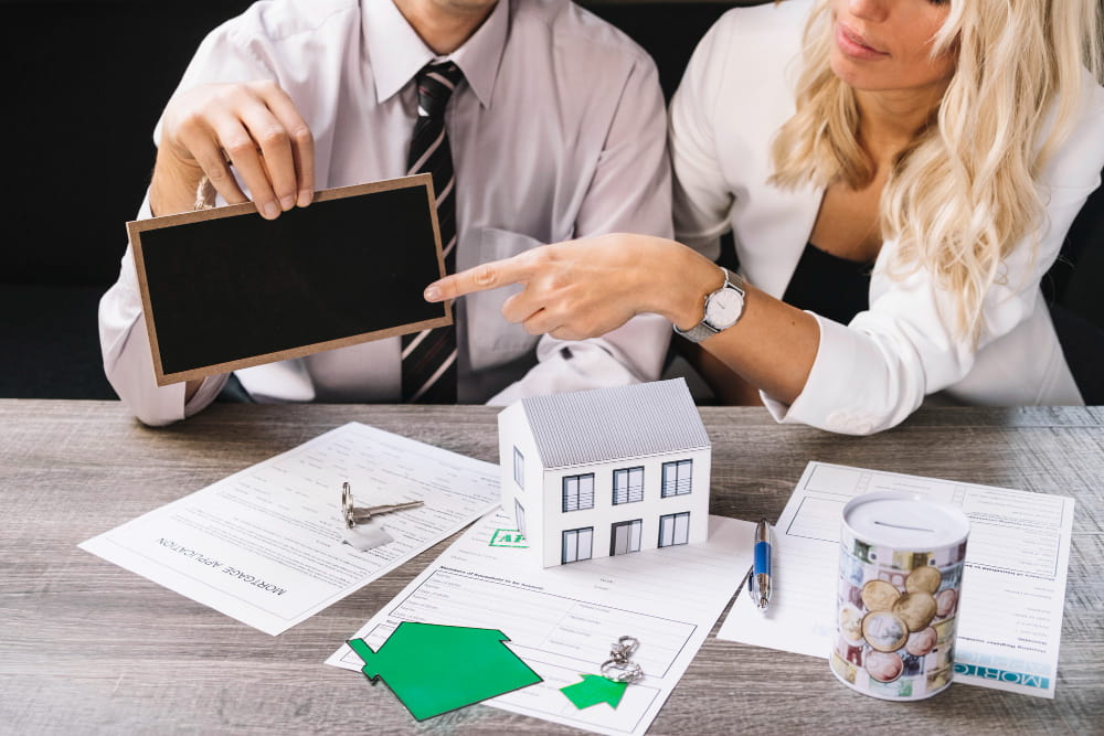 Two people sit at a table with real estate documents, a model house, a key, and a pot of coins.