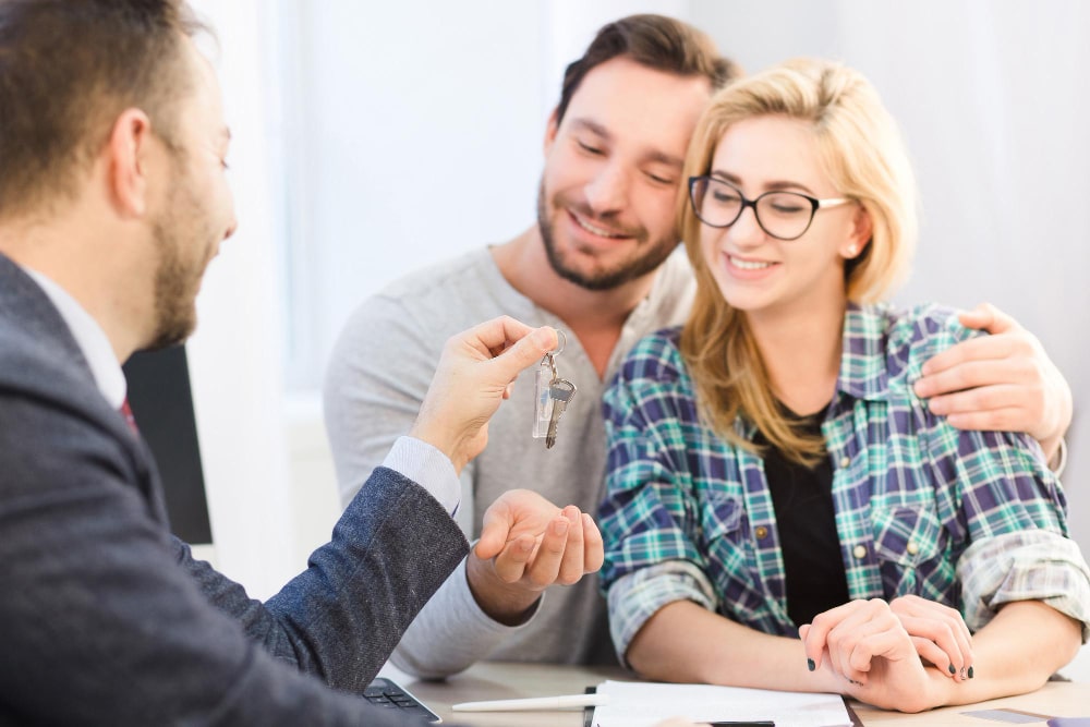 A couple smiles warmly as a businessman hands them keys, symbolizing a successful home purchase.