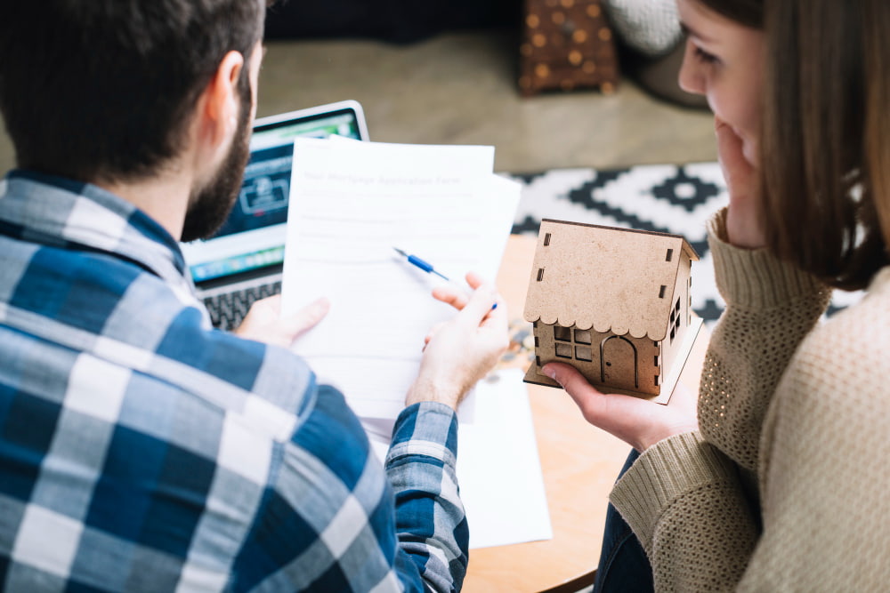 A couple discusses mortgage documents at a table. The man, in a plaid shirt, points at a paper. The woman, holding a house model.