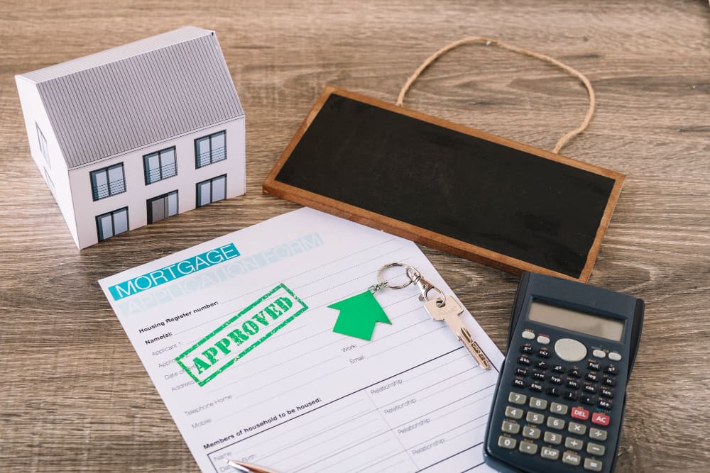 A miniature house model, a blank chalkboard, a calculator, a mortgage document stamped "Approved,”.