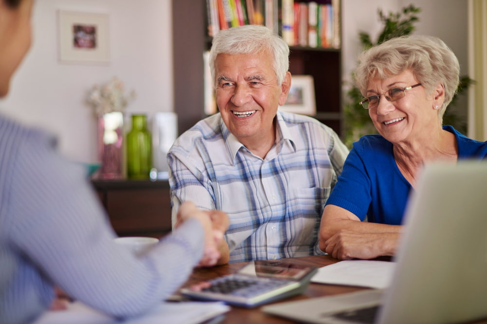Elderly couple at a table with a laptop, smiling as they shake hands with a person in the foreground.