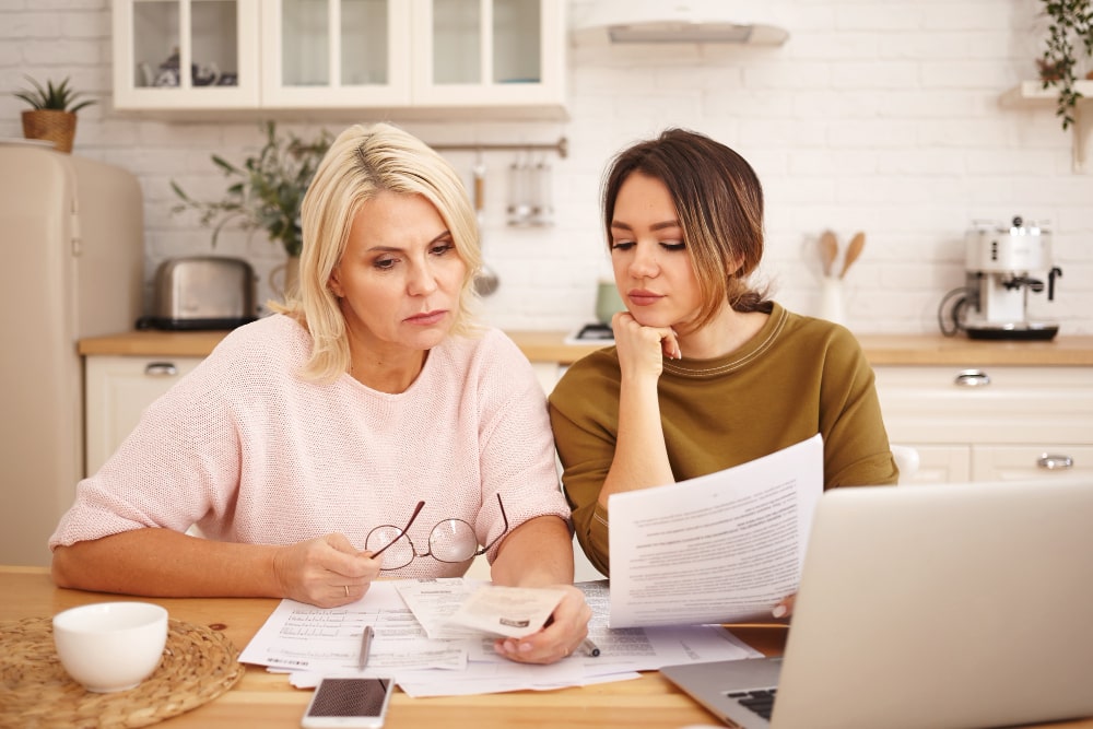 Two women sit at a kitchen table, focused on documents and a laptop.