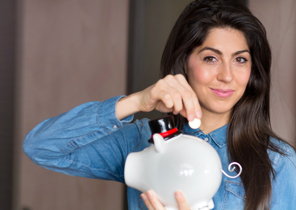 A woman in a denim shirt smiles while placing a coin into a piggy bank, symbolizing saving money.