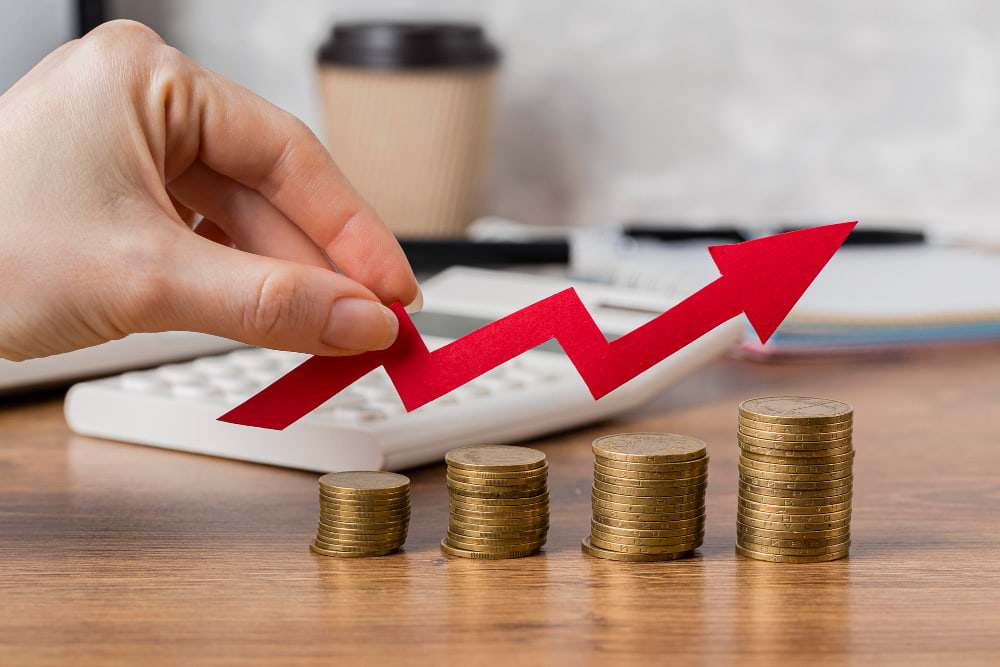 Hand holds red upward arrow above rising coin stacks—calculator and coffee cup blurred in back, symbolizing mortgage growth.