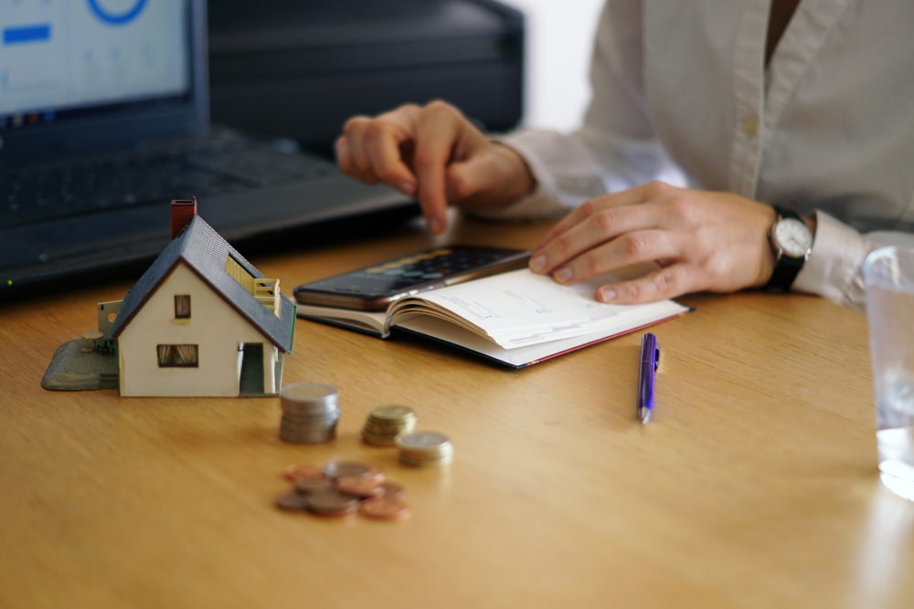 A person calculates finances at a desk with a small house model, coins, a notepad, calculator, and laptop.