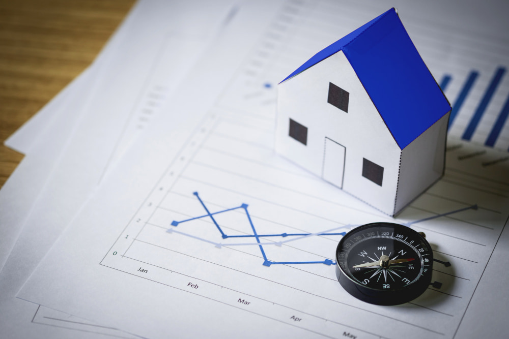 A paper house with a blue roof sits on financial graphs, alongside a compass.