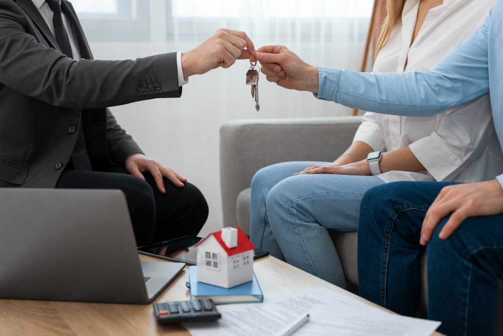 The agent hands keys to couple—laptop, papers, and house model on the table suggest a mortgage signing and new home purchase.