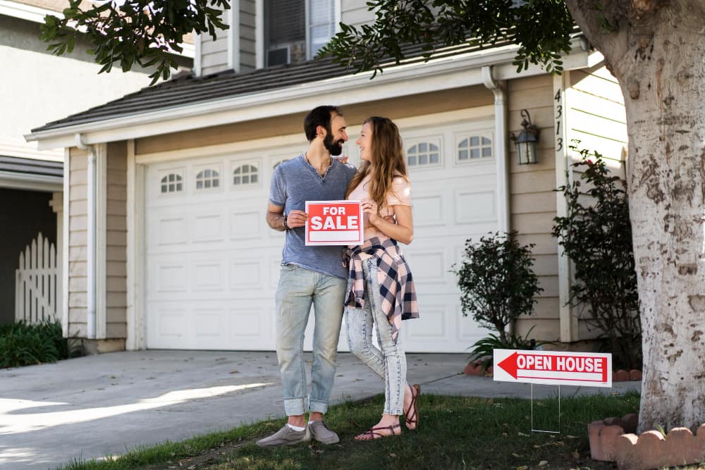 A smiling couple stands together in front of a house with a "For Sale" sign.