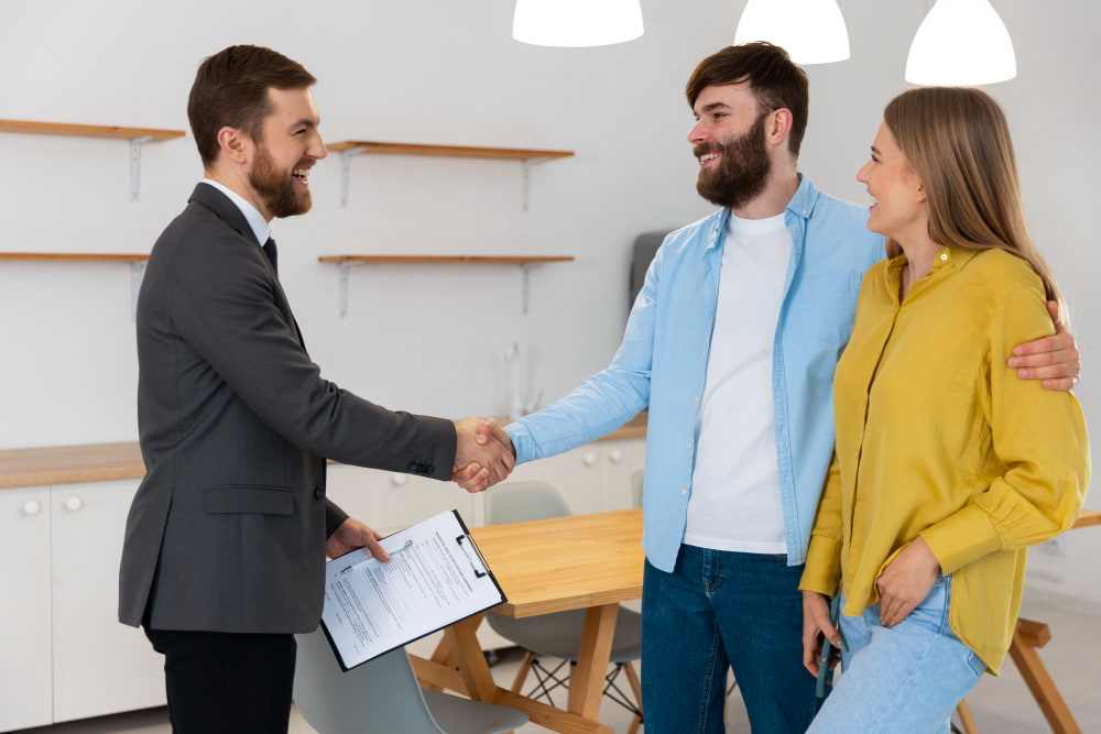 Smiling man in a suit shakes hands with another man while a woman in yellow stands in a bright mortgage meeting room.