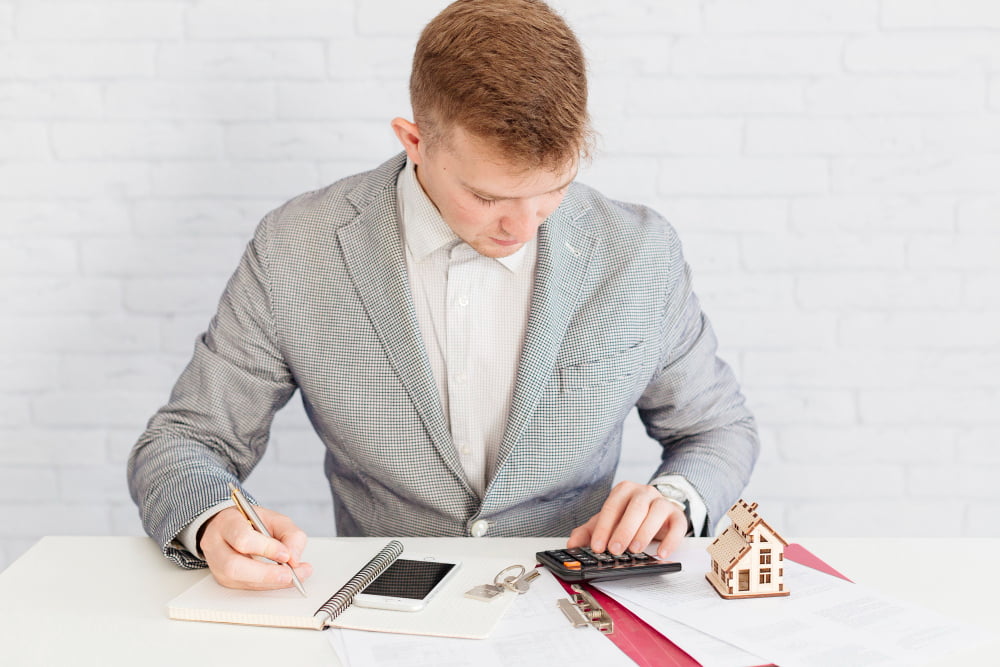 A man in a suit calculates with a calculator, surrounded by documents, a miniature house model, and a smartphone.