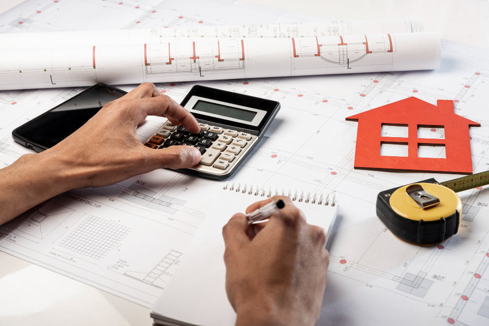 Hands with calculator and pencil over blueprints—smartphone, red house cutout, and tape measure suggest mortgage planning.