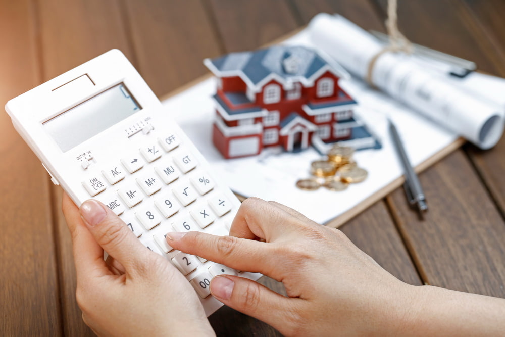 Hands using a calculator on a table with a model house, rolled-up blueprints, and coins in the background.