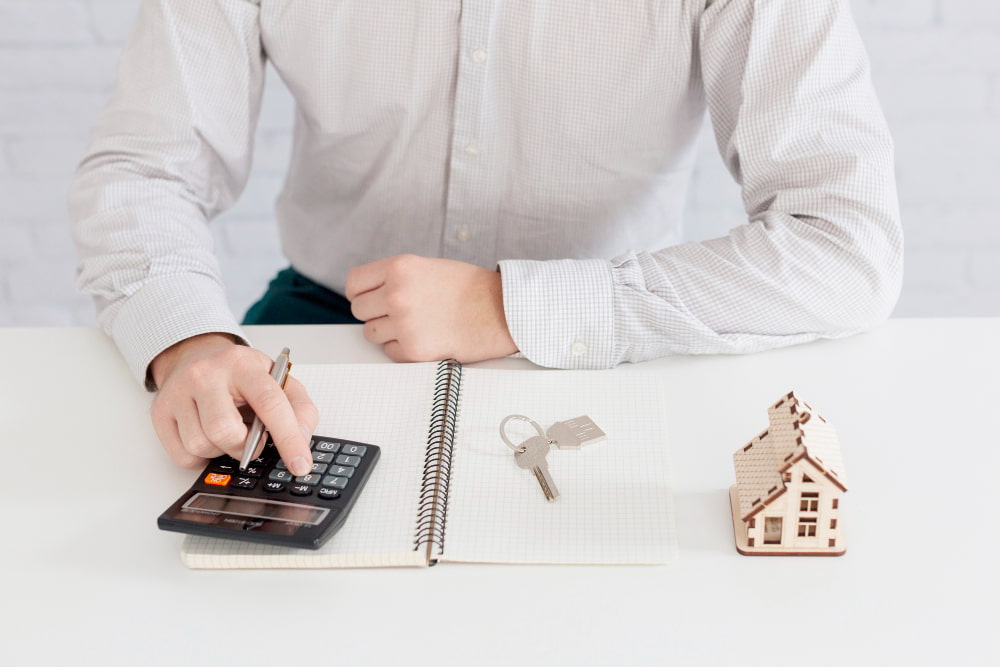 Person in white shirt calculates with pen and notebook—house model and keys suggest mortgage and real estate planning.