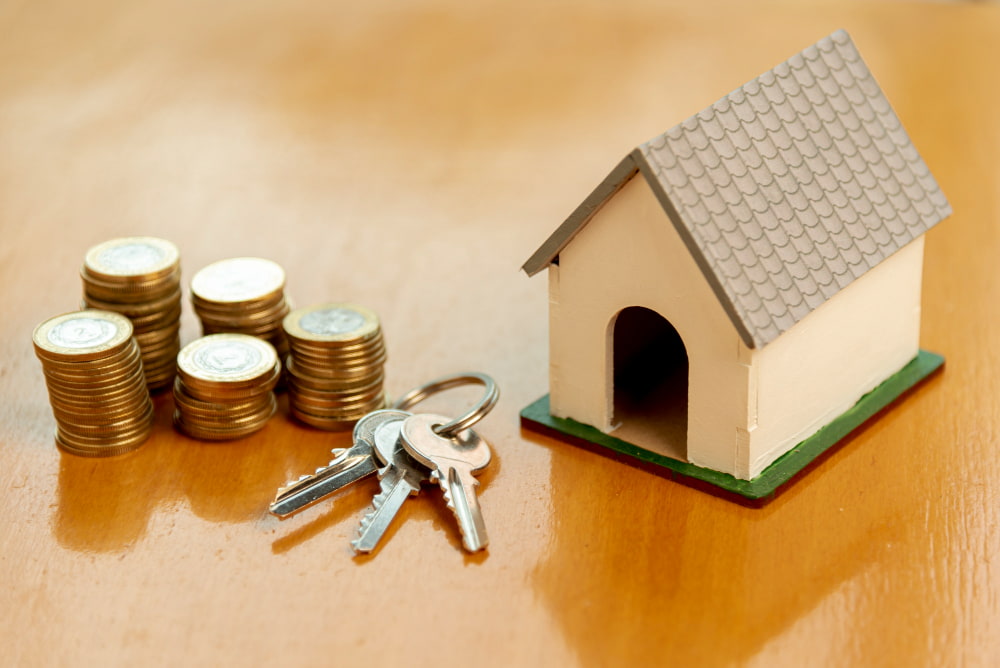 Stacks of gold coins, a set of keys, and a small model house sit on a wooden surface.