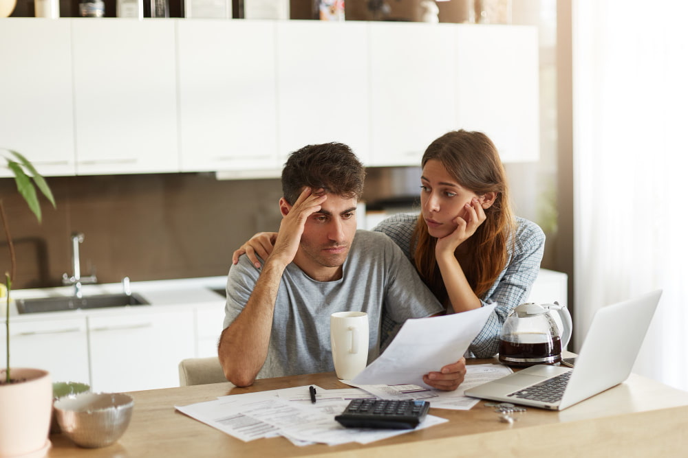 A concerned couple sits at a kitchen table, reviewing bills. The man holds his forehead, while the woman leans in supportively.
