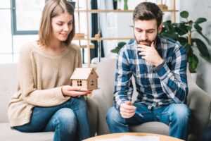 A couple sits on a couch in a bright room. The woman holds a model house, smiling, while the man looks thoughtful.