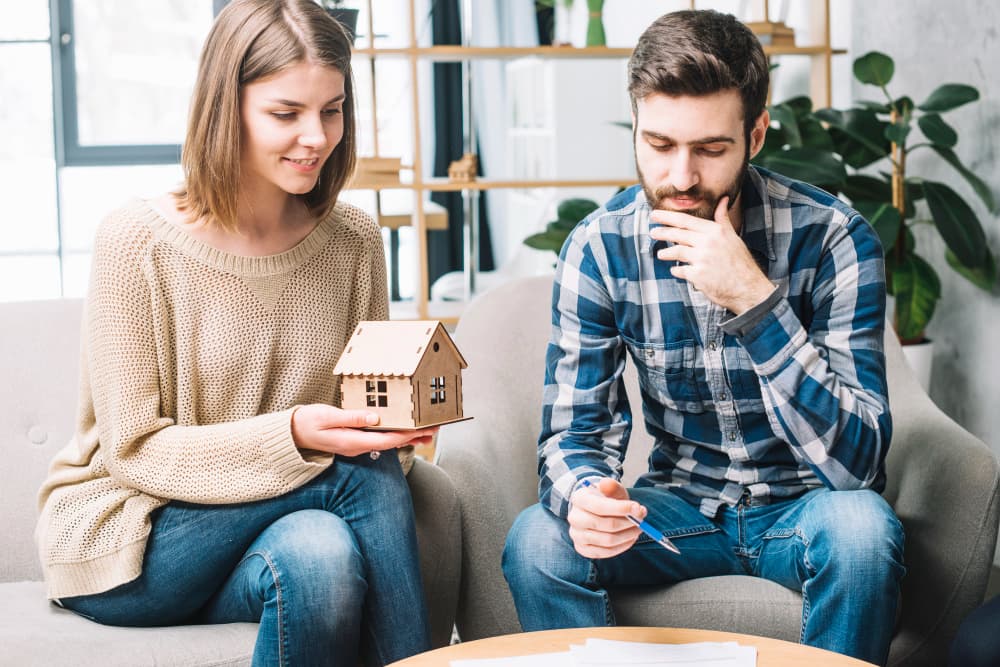A couple sits on a couch in a bright room. The woman holds a model house, smiling, while the man looks thoughtful.
