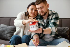 A couple sits on a couch holding a miniature house, symbolizing homeownership. They appear thoughtful and focused.