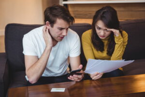 A worried couple sits on a sofa; the man holds a phone and the woman reads papers. They both look stressed.