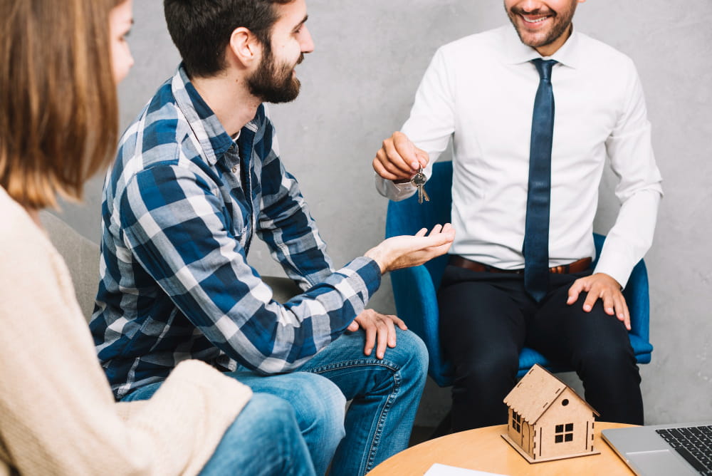 A smiling man in a shirt and tie hands house keys to a seated couple, symbolizing a new home.