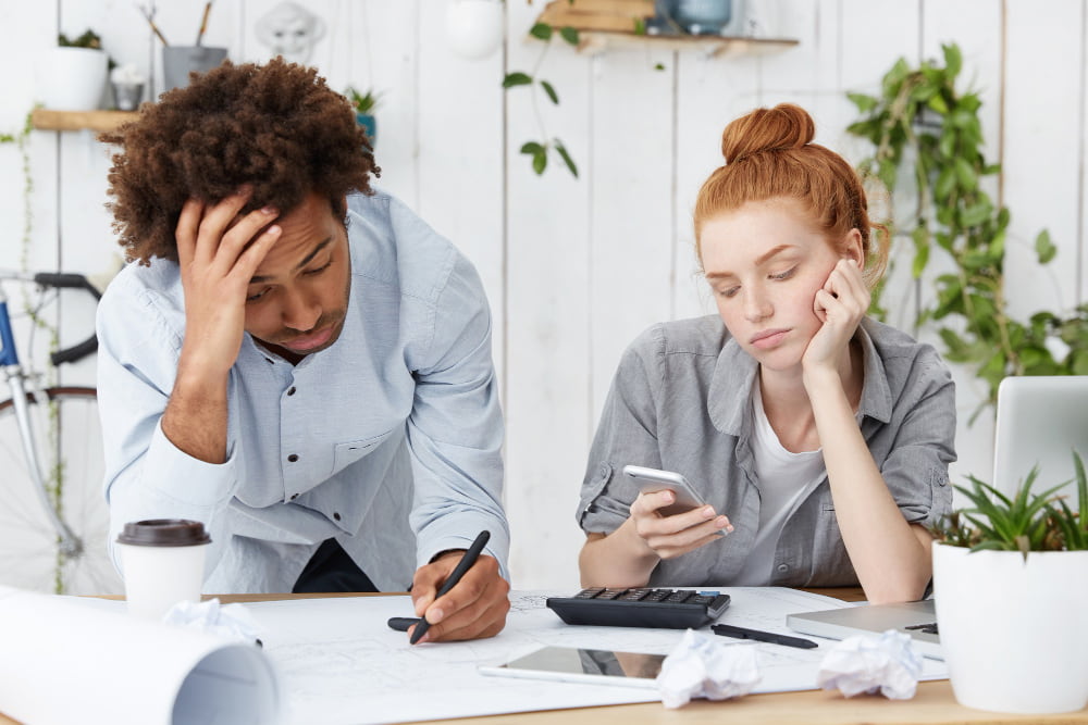 Frustrated pair at cluttered desk with papers, calculator, and laptop—woman checks phone, man writes, suggesting mortgage stress.