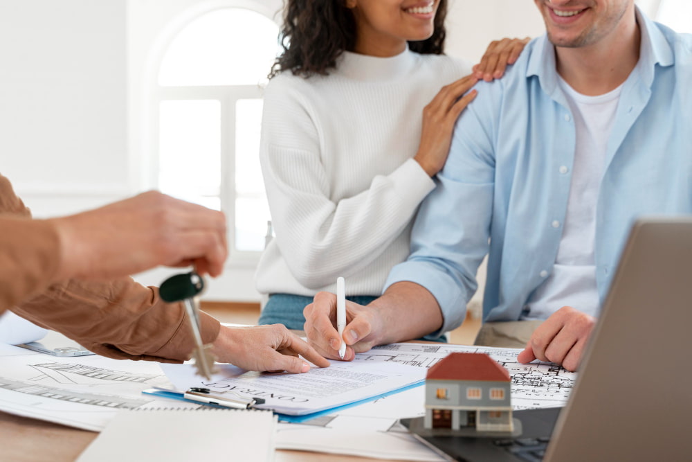 Couple smiling as they receive house keys from an agent, sitting by house plans and a laptop.